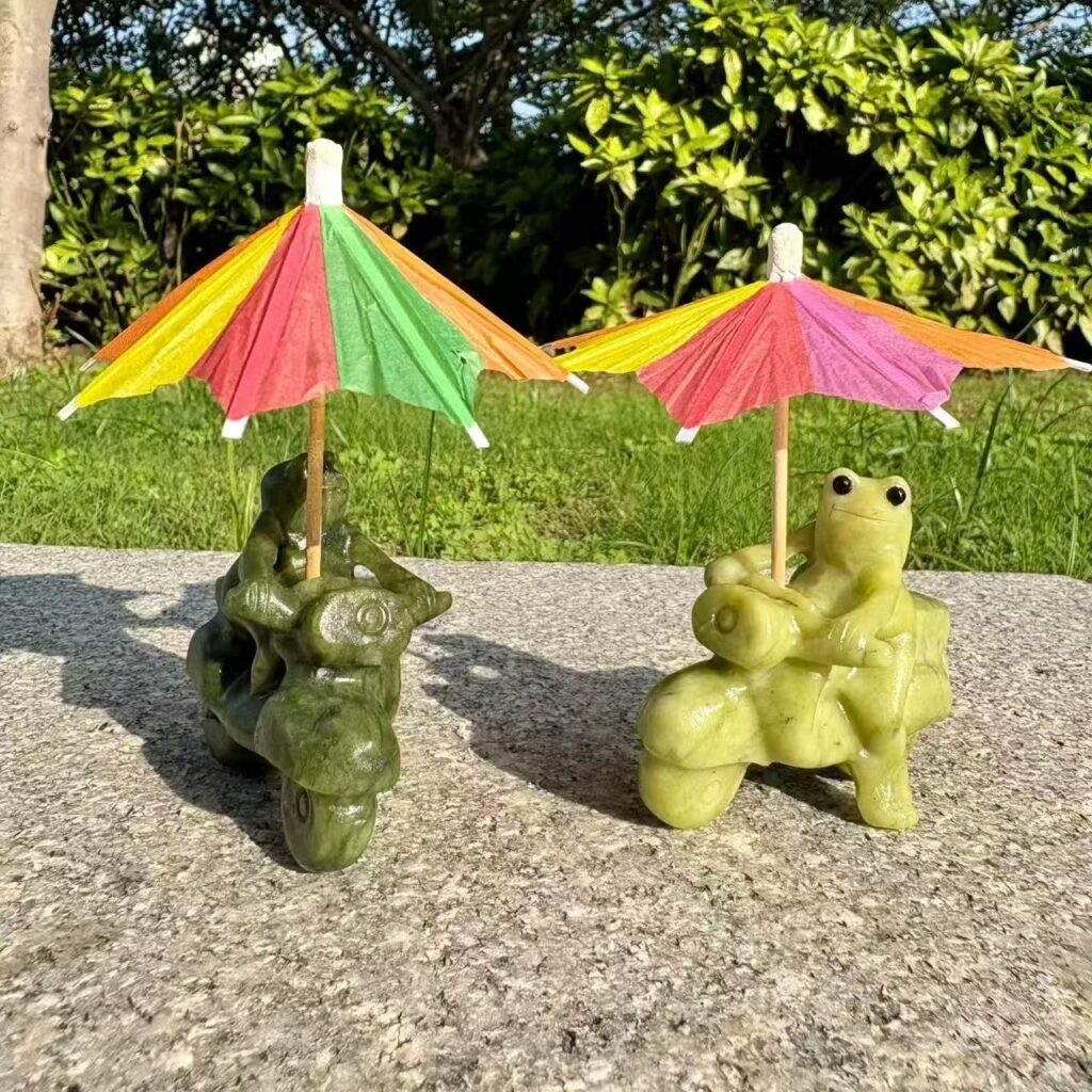 Two different handmade crystal frogs riding motorcycles, one light olive green and one deep forest green, posed with tiny colorful paper umbrellas.